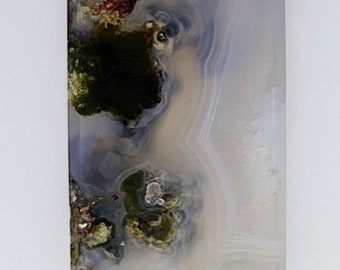 May include: A polished agate slab with a translucent, light blue background and green and brown inclusions. The agate has a natural, organic pattern.