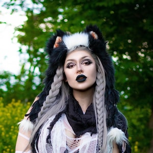 Peut inclure: Une femme portant un costume à capuche en fourrure noir et blanc avec un haut en maille blanche et un pantalon noir. Elle tient une seule rose blanche dans sa main et se tient dans un champ de fleurs jaunes.