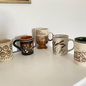 Può includere: Cinque tazze in ceramica di vari disegni e dimensioni sono disposte su una superficie bianca. Le tazze presentano motivi come vortici, motivi floreali e spighe di grano. Una tazza ha un'illustrazione di auto d'epoca.