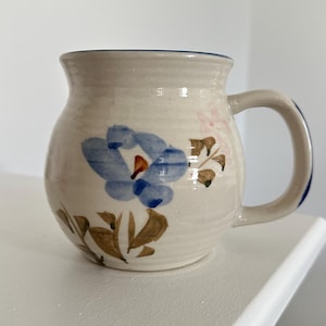 May include: Cream ceramic mug with a blue floral design. The mug has a rounded body, a curved handle, and a blue rim. Brown and pink accents complement the blue flower. The mug is likely handmade and suitable for hot drinks.