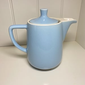 May include: A pale blue ceramic teapot with a matching lid and a white rim. The teapot has a curved handle and a spout for pouring. The teapot is a light blue colour and is on a white surface.