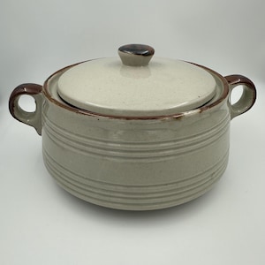 May include: A beige ceramic casserole dish with a lid. The dish has two handles and a brown rim. The lid has a small, rounded knob. The dish has horizontal, ridged detailing around the body.