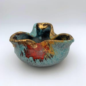 May include: A small, blue ceramic bowl with a textured surface. The bowl has splashes of red and gold paint and a slightly irregular shape.