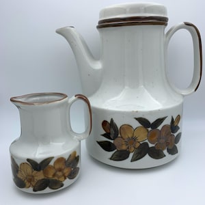 Könnte beinhalten: Ein weißes Keramik-Teekannen- und Milchkännchen-Set mit Blumenmuster. Die Teekanne hat einen braunen Deckel und einen braunen Griff. Das Milchkännchen hat einen braunen Griff. Die Blumen sind gelb und braun mit grünen Blättern.