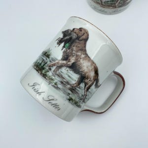 Könnte beinhalten: Weißer Keramikbecher mit braunem Henkel und Rand, der einen Irish Setter mit einem Vogel zeigt. Der Becher trägt die Aufschrift "Irish Setter". Die Illustration zeigt den Hund in einer natürlichen Umgebung, mit detaillierten Farben.