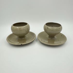 Könnte beinhalten: Zwei handgefertigte Keramikbecher mit angebrachten Untertassen. Die Becher sind rund mit einer kleinen Öffnung, und die Untertassen sind flach und kreisförmig. Die Keramik hat eine gesprenkelte beige Farbe.