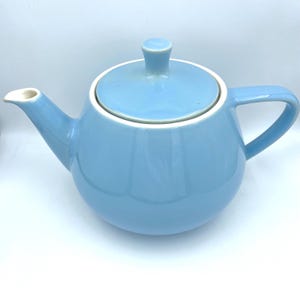 Può includere: Teiera in ceramica azzurra con coperchio e manico coordinati. La teiera ha un corpo arrotondato, un beccuccio curvo e un bordo bianco. Il coperchio ha un piccolo pomello. Progettata per preparare il tè.