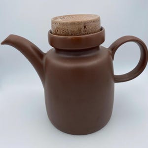 Könnte beinhalten: Eine braune Teekanne aus Keramik mit Deckel. Die Teekanne hat einen runden Körper und einen Ausguss auf einer Seite. Der Deckel ist aus hellbraunem Material und hat eine strukturierte Oberfläche.