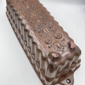 May include: A brown ceramic loaf tin with a decorative floral and scroll design. The rectangular tin has a ridged exterior and a hole on one end. The tin is approximately 30 cm long.