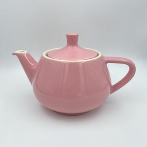 Könnte beinhalten: Eine rosa Keramik-Teekanne mit passendem Deckel. Die Teekanne hat einen abgerundeten Körper mit vertikalen Rippen, einen gebogenen Ausguss und einen Griff. Der Deckel hat einen kleinen Knopf zum einfachen Entfernen. Der Innenrand ist weiß.