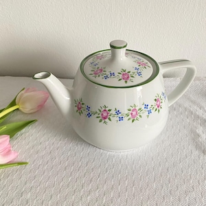 May include: White ceramic teapot with a green rim and handle. The teapot is decorated with pink and blue floral patterns. A matching lid sits on top. A pink tulip is in the background.