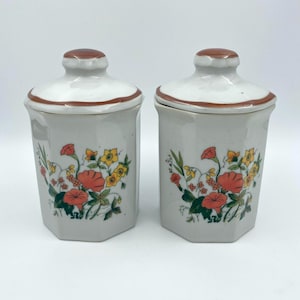May include: A pair of ceramic storage jars with lids. The jars are light grey with a floral pattern in orange, yellow, and green. The lids are white with a brown trim and a small knob. The jars are hexagonal in shape.