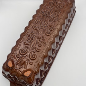 Könnte beinhalten: Eine braune Keramik-Kastenform mit glänzendem Finish. Die Form hat ein dekoratives Blumen- und Wirbelmuster auf der Oberseite und ein geriffeltes Design an den Seiten. Die Form hat einen Griff mit einem Loch zum Aufhängen.