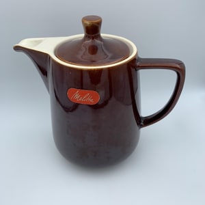 Könnte beinhalten: Eine braune Kaffeekanne aus Keramik mit einem weißen Deckel und einem roten Etikett mit der Aufschrift "Melitta".