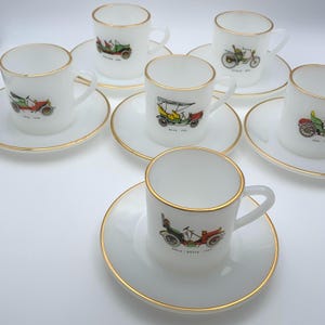 Könnte beinhalten: Satz mit sechs weißen Porzellan-Teetassen und Untertassen mit goldener Verzierung. Jede Tasse zeigt eine andere Vintage-Auto-Illustration mit der Marke und dem Jahr, die auf dem Boden aufgedruckt sind.