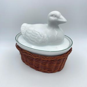 Op de afbeelding: Een witte keramische eendvormige schaal met een deksel, geplaatst op een witte ovale schaal met een groene rand. De schaal staat in een geweven bruine mand. De eend heeft gedetailleerde veren en een gebogen snavel.