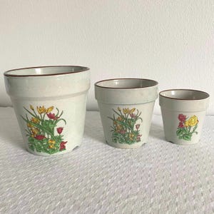 Könnte beinhalten: Set aus drei Keramik-Blumentöpfen. Die Töpfe sind cremefarben mit einer gesprenkelten Textur und einem braunen Rand. Jeder Topf hat ein Blumendesign mit gelben, roten und grünen Blumen. Der größte Topf hat einen Durchmesser von ca. 18 cm.