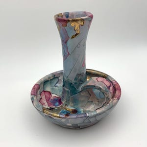 Op de afbeelding: Een keramische kandelaar met een kleurrijk, mozaïekachtig ontwerp. De basis is een ondiepe schaal met een verhoogde, gebogen handgreep. De kandelaar is versierd met tinten blauw, roze en goud.