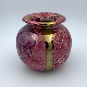 May include: A small, round ceramic vase with a crackled glaze in shades of pink and purple. The vase has a gold stripe down the side.