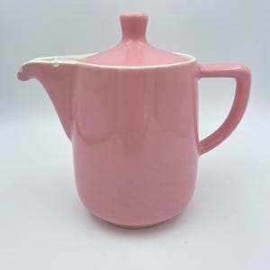 May include: A pink ceramic creamer with a matching lid. The creamer has a spout, a handle, and a white rim. The lid has a small knob on top. The creamer is a solid, glossy pink colour.
