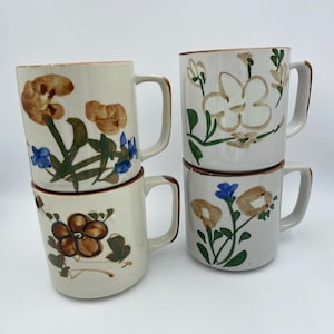 Op de afbeelding: Vier gestapelde keramische mokken met bloemmotieven. De mokken zijn gebroken wit met bruine, blauwe en groene geschilderde bloemen. Elke mok heeft een bruine rand en een handvat.