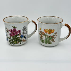 Op de afbeelding: Twee witte keramische mokken met een gespikkelde afwerking en bruine randen. De linker mok toont een boeket roze en blauwe wilde bloemen. De rechter mok heeft oranje lelies en groen blad, beide voorzien van een gebogen handvat.