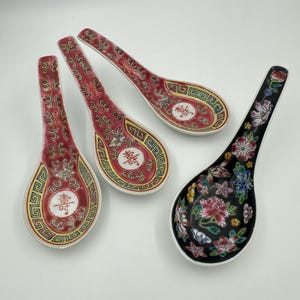 May include: Four ceramic Chinese soup spoons. Three spoons have a red background with ornate gold and white designs, and Chinese characters. One spoon has a black background with a floral design in pink, blue, and white.