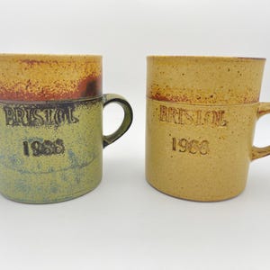 May include: Two ceramic mugs with handles. One is green, the other is tan. Both mugs have a band of brown and yellow at the top and the word "BRISTOL" and the year "1988" printed on the front.