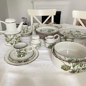 Peut inclure: Service de table complet en grès vintage avec une finition mouchetée blanche, un bord marron et un motif botanique vert. L'ensemble comprend une théière, des tasses, des soucoupes, des assiettes, des bols, une crémière et un sucrier.