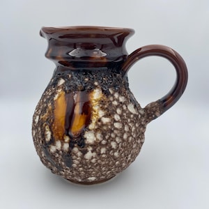 Könnte beinhalten: Ein brauner Keramikkrug mit einer strukturierten, gesprenkelten Oberfläche. Der Krug hat einen dunkelbraunen Henkel und Rand, mit einer helleren braunen Glasur in Bereichen. Das Design ist rustikal und vintage.