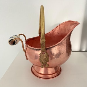 May include: A copper sugar scuttle with a brass handle and decorative elements. The scuttle features a spout and a small, ornate handle with a blue and white pattern. The base is round, and the handle is attached with a decorative lion's head.