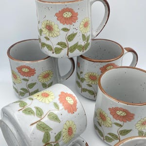 Può includere: Un set di sei tazze in ceramica con finitura bianca maculata. Ogni tazza presenta un motivo floreale con fiori arancioni e gialli, foglie verdi e dettagli marroni. Le tazze hanno un design con manico classico.
