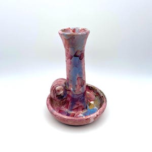 May include: A handmade ceramic candlestick holder with a distinctive, crackled glaze. The colour scheme features pinks, purples, and blues. The candlestick holder has a circular base, a tall, slender stem, and a small handle.