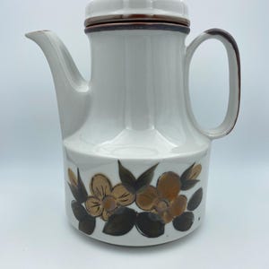 Könnte beinhalten: Weiße Kaffeekanne aus Keramik mit braunem Deckel und Blumendesign. Die Blumen sind gelb und braun mit grünen Blättern.