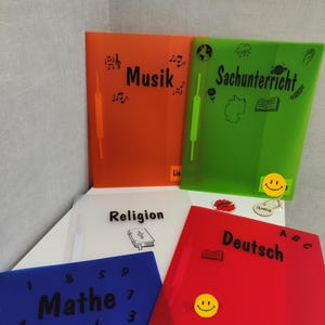 Könnte beinhalten: Fünf bunte Plastikordner mit verschiedenen Fächern bedruckt: Musik, Sachunterricht, Religion, Deutsch und Mathe. Jeder Ordner hat einen Smiley-Aufkleber.