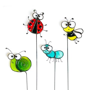 May include: Four whimsical garden stakes featuring insect designs. The set includes a ladybug with a red and black body, a yellow and black bee, a green snail, and a blue ant. Each stake has a metal rod for easy placement in the garden.