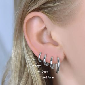 May include: Silver hoop earrings of varying sizes, shown on an ear. Sizes are 6mm, 8mm, 10mm, and 14mm. The earrings are shiny and reflect light. The ear is set against a background of blonde hair.