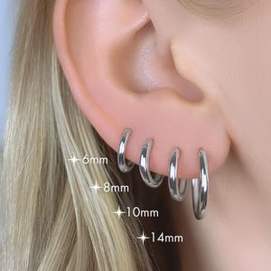 May include: An ear adorned with four silver hoop earrings, sized at 6mm, 8mm, 10mm, and 14mm. The earrings are highly polished, reflecting light. The background is softly blurred, focusing attention on the jewellery.