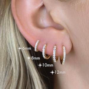 May include: Gold hoop earrings with clear, sparkling stones. The earrings are shown in four sizes: 6mm, 8mm, 10mm, and 12mm. The earrings are displayed on an ear, showcasing their size and style.