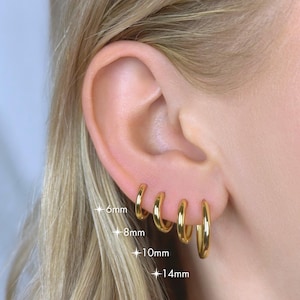 May include: A close-up of a person's ear adorned with four gold hoop earrings of varying sizes: 6mm, 8mm, 10mm, and 14mm. The earrings are shiny and reflect light, showcasing a classic jewelry style. The ear is set against a background of blonde hair.