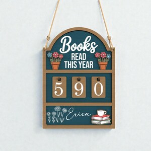 以下が含まれることがあります： 「Books Read This Year」の文字と「590」の数字が入った木製のサイン。ティール色の背景に、花の装飾と本のイラストが描かれています。「Erica」という名前も含まれており、読書進捗を追跡するように設計されています。