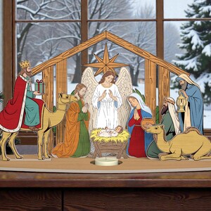 Puede incluir: Un belén de madera con recortes coloridos de la Sagrada Familia, los Reyes Magos, un ángel y el niño Jesús en un pesebre. La escena está ambientada frente a una ventana invernal, con una estrella sobre el establo.