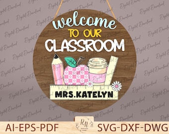 Puede incluir: Un letrero redondo de madera para el aula con las palabras "Welcome to Our Classroom" en blanco y azul claro. El letrero presenta un lápiz, una manzana, una taza de café, una regla y el nombre "Mrs. Katelyn".