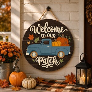 May include: A round dark wood sign hanging by a rope, featuring the text Welcome to our Patch in cream lettering. A blue vintage truck illustration carries orange pumpkins in its bed, accented by small orange and green autumn leaf cutouts.