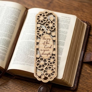 Może przedstawiać: Drewniana zakładka do książki z kwiatowym wzorem i napisem "The joy of the Lord is your strength" w owalu. Zakładka znajduje się w otwartej książce z brązową skórzaną oprawą.