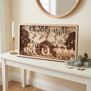 May include: A rectangular wooden nativity scene display featuring layered cutouts of the holy family, camels, sheep, and palm trees. The text "Oh Holy night" is carved into the top section, accented by stars and flying angels in a light and dark wood finish.
