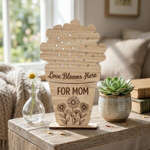 May include: Wooden floral-shaped sign with the text "Love Blooms Here FOR MOM" and daisy illustrations. The sign is on a wooden table next to a small vase with a daisy and a potted succulent.