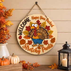 May include: A round wooden sign featuring a cartoon turkey wearing blue pants, surrounded by colorful autumn leaves. The text reads "WELCOME Hope You Packed Stretchy Pants" in black and orange lettering. The sign hangs from a brown rope.