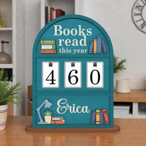 May include: A teal wooden book counter with the text "Books read this year" and the number "460". The name "Erica" is displayed below the counter, along with illustrations of books and a desk lamp.