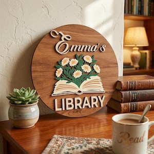 May include: A round wooden sign with the text "Emma's LIBRARY" in a layered design. The sign features a floral arrangement above an open book. The sign is brown with cream-colored lettering and accents.